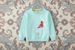 Light blue sweatshirt with floral design and embroidered character on a white background